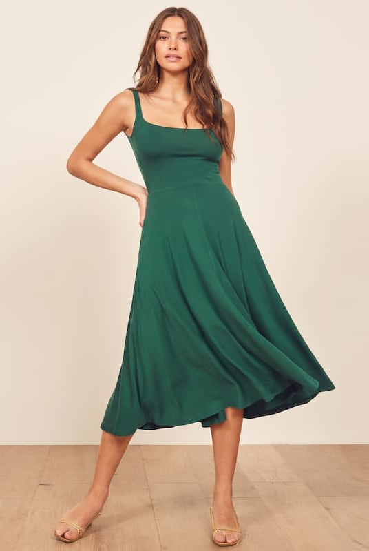 A woman modeling a sleeveless, scoop-neck midi dress in emerald green, standing on a light wood floor against a plain beige wall.