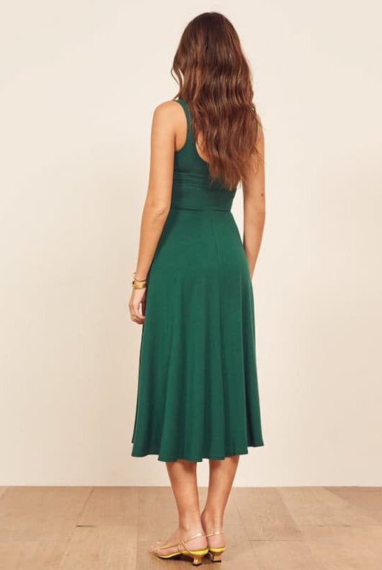 A woman viewed from behind wearing a sleeveless emerald green midi dress with a fitted bodice and flared skirt, standing against a plain beige wall on a light wood floor.