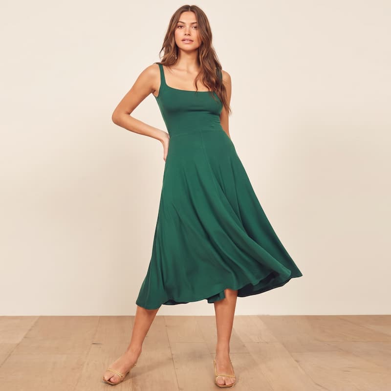 A woman modeling a sleeveless, scoop-neck midi dress in emerald green, standing on a light wood floor against a plain beige wall.