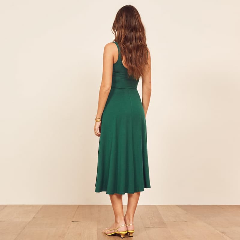 A woman viewed from behind wearing a sleeveless emerald green midi dress with a fitted bodice and flared skirt, standing against a plain beige wall on a light wood floor.