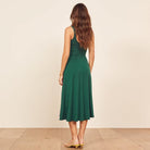 A woman viewed from behind wearing a sleeveless emerald green midi dress with a fitted bodice and flared skirt, standing against a plain beige wall on a light wood floor.