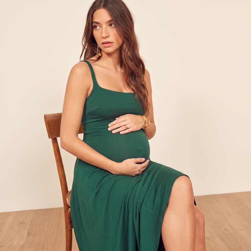 A pregnant woman wearing a dark green sleeveless maternity dress sits on a wooden chair against a plain beige wall.