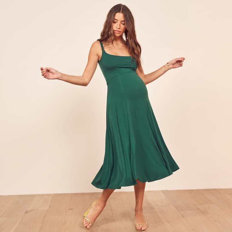 A woman modeling a sleeveless, scoop-neck midi dress in emerald green, standing against a light beige wall and light wood floor.