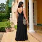A woman wearing a black maxi dress with thin straps and a low open back, standing on a tiled walkway in front of a building with white columns. She is carrying a taupe crossbody bag.