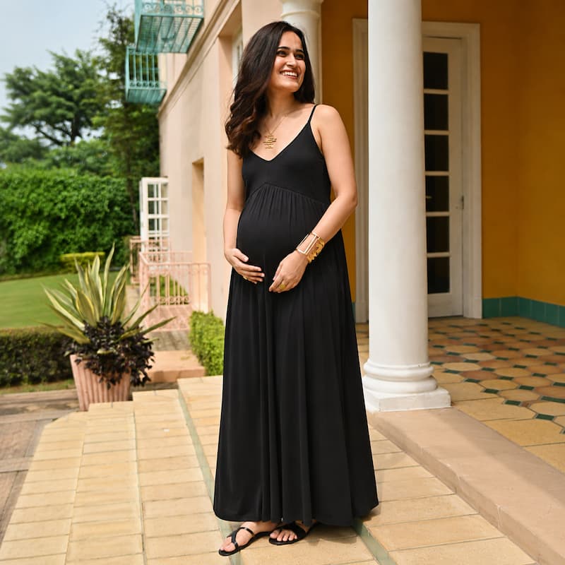 A pregnant woman wearing a black maternity maxi dress with spaghetti straps and a V-neckline, standing on a tiled walkway in front of a building with white columns.